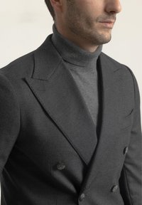 Blazer de charco negro de doble botonadura con solapa muesca, combinado con un cuello de tortuga gris ajustado. Textura suave con botones negros y un detalle mínimo.
