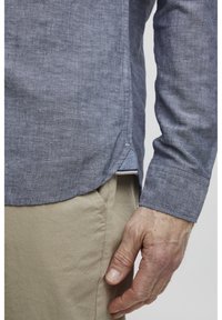 Camicia di lino a maniche lunghe, blu chiaro, con tessuto testurizzato, orlo curvo e polsini con bottoni, abbinata a pantaloni di cotone color beige.