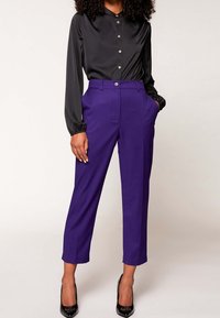 Kvinna iklädd en svart satin knäppt blus, högmidjade lila capribyxor och svarta spetsiga klackskor, stående med händerna i fickorna.