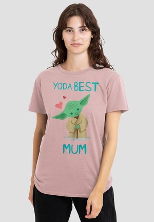 Frau mit langen braunen Haaren, die ein hellrosa T-Shirt mit einem Cartoon-Yoda, Herzen und dem Text "Yoda Beste Mama" auf grauem Hintergrund trägt.
