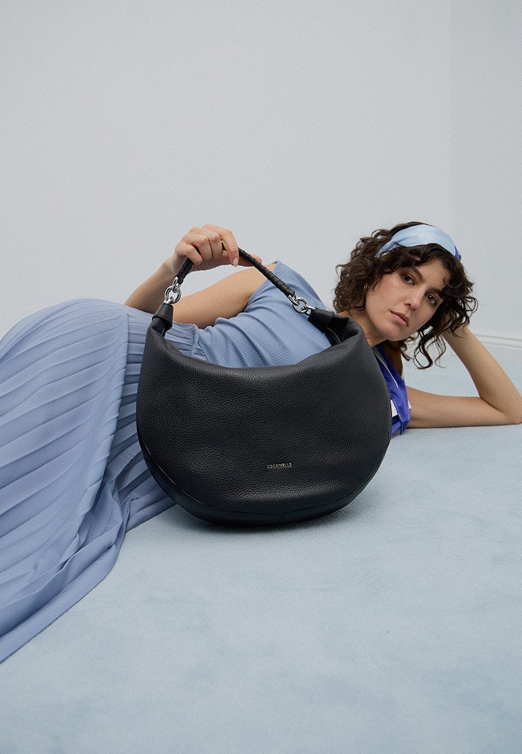 Donna in abito azzurro chiaro e fascia per capelli è sdraiata sul pavimento, mentre tiene in mano una grande borsa in pelle nera con chiusure metalliche.