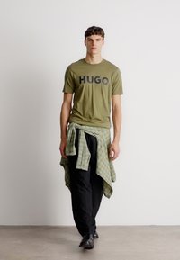 Olivovo zelené bavlnené tričko s nápisom "HUGO" v čiernej farbe, doplnené čiernymi nohavicami a zelenou kockovanou košeľou zviazanou okolo pásu.