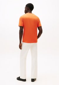Πορτοκαλί βαμβακερό μπλουζάκι με κοντά μανίκια, στρογγυλή λαιμόκοψη και λεία υφή, συνδυασμένο με άσπρο παντελόνι φαρδιάς γραμμής και μαύρα παπούτσια.