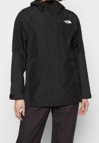 Czarna wodoodporna kurtka z kapturem, kieszeniami na zamek i detalem z logo. Materiał jest gładki o matowym wykończeniu.