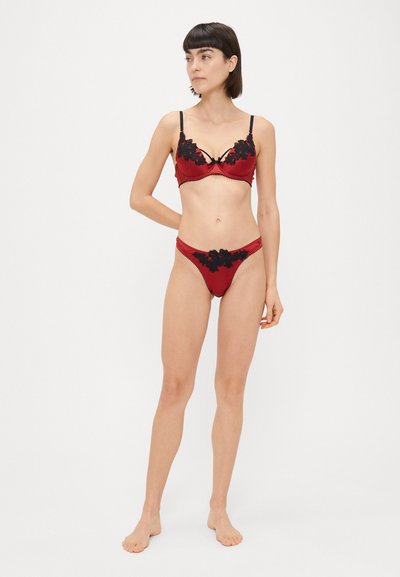 Červený saténový spodný prádlo so čiernymi čipkovými detailmi. Podprsenka má nastaviteľné ramienka a kosticu; tangá majú podobný čipkový detail.