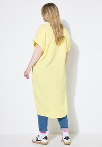 Vestido amarillo, de gran tamaño, con mangas cortas y una tela texturizada, combinado con jeans azules y zapatillas en blanco y negro con acentos de calcetines rosas.
