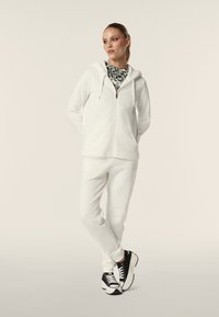 Felpa bianca con zip e un interno morbido, abbinata a pantaloni della tuta bianchi. Sneakers bianche e nere completano l'outfit.