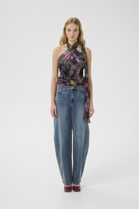 Donna in piedi che indossa un top a fascia colorato con un lungo drappo, jeans blu a vita alta e a gamba larga, e scarpe burgundy con punta quadrata.
