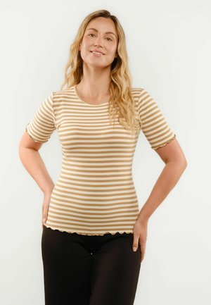 Kvinde med langt bølget blondt hår iført en beige og hvid stribet kortærmet top og sorte bukser, stående med hænderne på hofterne.