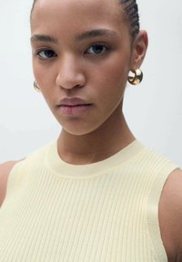 Pletivový top bez rukávov v svetložltej farbe s rebrovanou textúrou. Model má na sebe lesklé zlaté kruhové náušnice. Neutrálny pozadie.