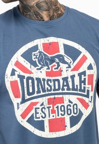 Mėlynas medvilninis marškinėliai su nublukusiu apskritu logotipu, kuriame pavaizduotas liūtas, raudonais ir baltais akcentais, ir užrašu "Lonsdale EST. 1960".