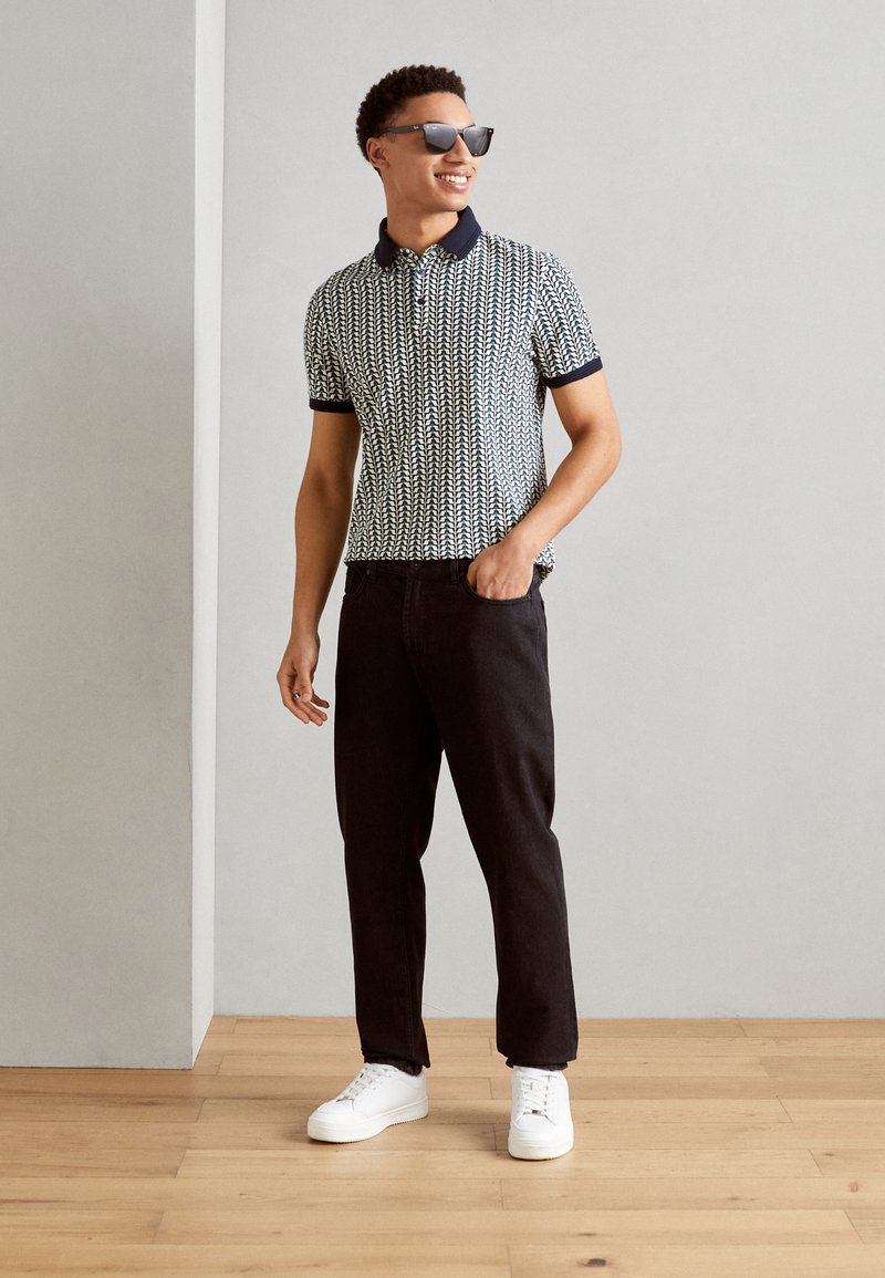 Kurzärmliges Polohemd mit navyfarbenem Kragen und einem gemusterten Design in Hellblau und Weiß. Tragend mit dunklen Jeans und weißen Sneakern.