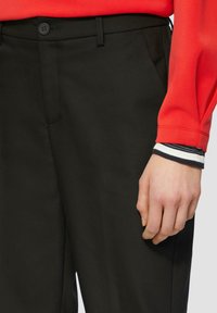 Zwarte op maat gemaakte broek met een gladde textuur en voorzakken, gecombineerd met een rode blouse met lange mouwen en gestreepte manchetten.
