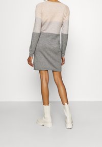 Person som bär en långärmad, färgblockerad klänning i beige, grått och kolgrått, i kombination med vita ankelstövlar, vänd bort på en ren bakgrund.