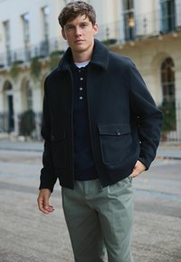 Nuori mies ulkona musta vetoketjullinen takki, jossa suuret taskut, päällä tumma paita ja vaaleanharmaat housut, taustalla rakennuksia.