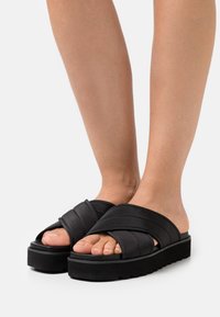 Sandali neri con cinturini incrociati imbottiti e una suola spessa e texture. Presenta una piattaforma elevata per un comfort aggiuntivo.