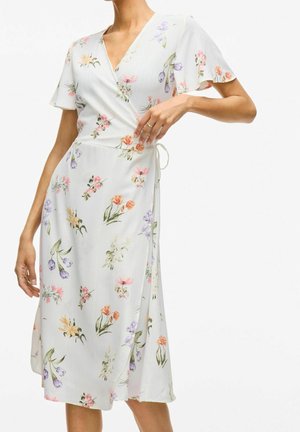 Robe portefeuille blanche avec manches courtes volantées et imprimé floral multicolore, portée par un mannequin avec la main sur la taille montrant le détail du lien.