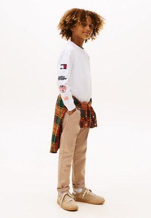 Langärmliges weißes Hemd mit Logo und Patches, beige Hose und ein kariertes Hemd um die Taille gebunden. Braune Sneaker mit weißen Sohlen.