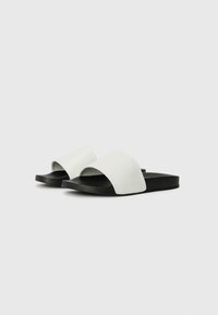 Hvide slip-on sandaler med en struktureret sort sål, der har en bred rem og et åbent tå-design. Glat syntetisk materiale.