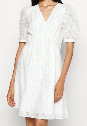 Femme portant une robe blanche avec des manches bouffantes mi-longues, un col en V et un tissu texturé à rayures horizontales.