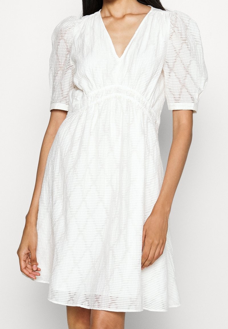 Femme portant une robe blanche avec des manches bouffantes mi-longues, un col en V et un tissu texturé à rayures horizontales.