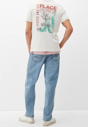 Persoon draagt een wit T-shirt met rode en groene Palm Beach grafische tekst en een blauwe spijkerbroek, staand tegen een eenvoudige witte achtergrond.