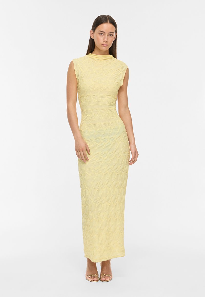 Femme portant une robe maxi sans manches, texturée, jaune pâle avec un col montant, debout les mains relâchées le long du corps sur un fond blanc.