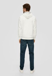 Un sweat à capuche blanc avec un cordon de serrage, portant un texte dans le dos, assorti à un jean bleu foncé et des baskets blanches.