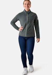 Grå zip-up fleecejacka med hög krage, texturerat tyg och logotyp på ärmen, kombinerad med mörka jeans och vita sneakers.