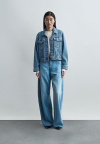 Γυναίκα όρθια, φοράει μπλε τζιν τζάκετ, τζιν παντελόνι με ψηλή μέση και φαρδιά μπατζάκια, λευκή ριμπ μπλούζα και μαύρα παπούτσια, μπροστά σε απλό φόντο.