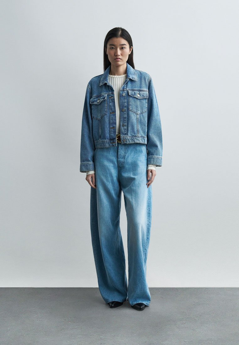 Femme debout portant une veste en denim bleue, un jean taille haute à jambes larges, un haut blanc côtelé et des chaussures noires, sur un fond uni.