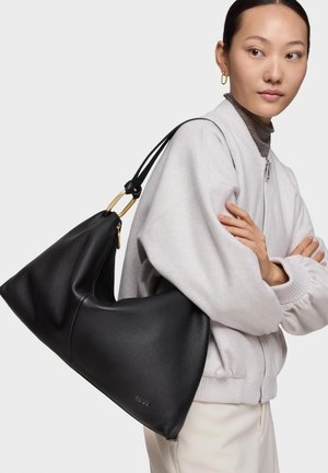 Donna che indossa una giacca leggera e un dolcevita, che porta una grande borsa a tracolla in pelle nera con dettaglio di fibbia dorata.