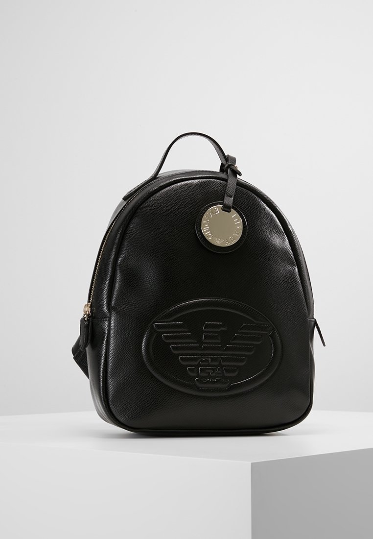 Sac à dos en cuir noir à forme arrondie, avec un logo en relief sur le devant et une étiquette circulaire argentée. Comprend une poignée supérieure et des bretelles ajustables.