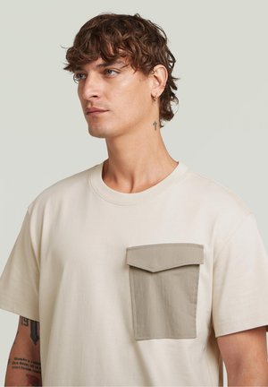 T-shirt beige in cotone con tasca applicata grigia a contrasto, scollo rotondo e maniche corte, dal texture liscia e vestibilità comoda.