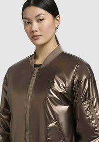 Metalická hnědá bomber bunda se zipem, ribbením límcem, bočními kapsami a logem na hrudi. Hladká, lesklá textura.