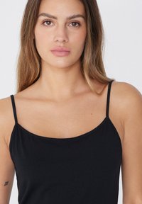 Černý top na ramínka s tenkými popruhy, který má hladkou texturu a přiléhavý střih. Bez výrazných vzorů nebo akcentů.