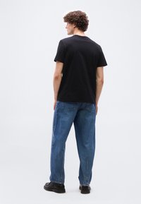 Άνδρας με σγουρά μαλλιά, φοράει μαύρη μπλούζα, μπλε τζιν και μαύρα παπούτσια, στέκεται και κοιτάζει προς τα αριστερά μπροστά σε ένα απλό φόντο.