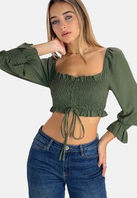 Grøn cropped bluse med smocket tekstur, firkantet halsudskæring og flagrende ærmer, parret med blå jeans. Materialet ser letvægtigt ud.