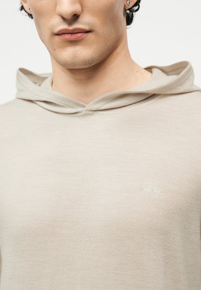 Άνδρας που φοράει μπεζ πλεκτή κουκούλα με ένα μικρό κεντημένο λογότυπο στο στήθος, εμφανίζοντας λεπτομέρειες του λαιμού και του κάτω μέρους του προσώπου.