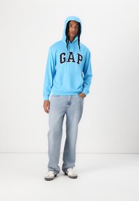 Light blue hoodie with "GAP" in dark navy, featuring a front pocket and a drawstring hood, paired with light blue jeans and black-and-white sneakers.