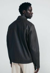 Giacca di cuoio marrone con colletto strutturato, polsini con bottoni e una texture liscia, esposta da un'angolazione laterale che mette in evidenza la vista posteriore.