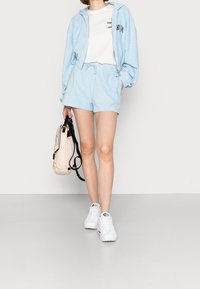 Light blue zip-up hoodie and matching shorts in soft fabric, paired with a white t-shirt, white sneakers, and a beige backpack.