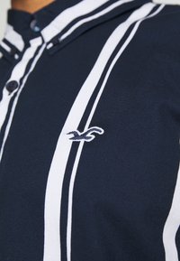 Mörkblå skjorta med vertikala vita ränder, knappstängning och en broderad logotyp på vänster sida nära kragen.