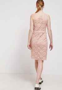 Kant jurk in lichtroze met een asymmetrisch ontwerp, gestructureerde stof, getailleerde silhouet en geschulpte zoom. Draagt met witte schoenen.