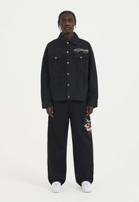 Giubbotto di jeans nero con due tasche sul petto, bottoni argentati e logo ricamato bianco. Abbinato a pantaloni neri larghi con ricami colorati.