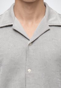 Gros plan d’une personne portant une chemise à fines rayures gris et blanc avec un col relax et des boutons blancs.
