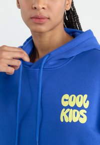 Blue hoodie made of soft fabric with a drawstring. Features a large yellow "COOL KIDS" graphic on the front. Smooth texture, casual design.
