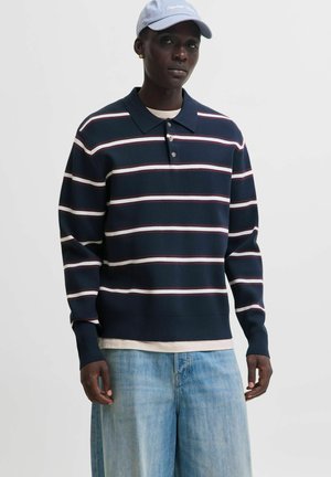 Modello maschio di colore che indossa una polo a righe blu navy, jeans azzurri e un berretto da baseball azzurro, in piedi davanti a uno sfondo semplice.