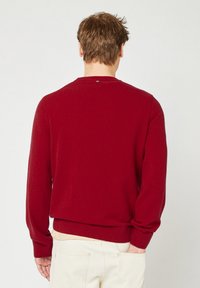 Uomo con capelli corti castani che indossa un maglione rosso e pantaloni beige chiaro, in piedi con una mano in tasca e la schiena rivolta in avanti.