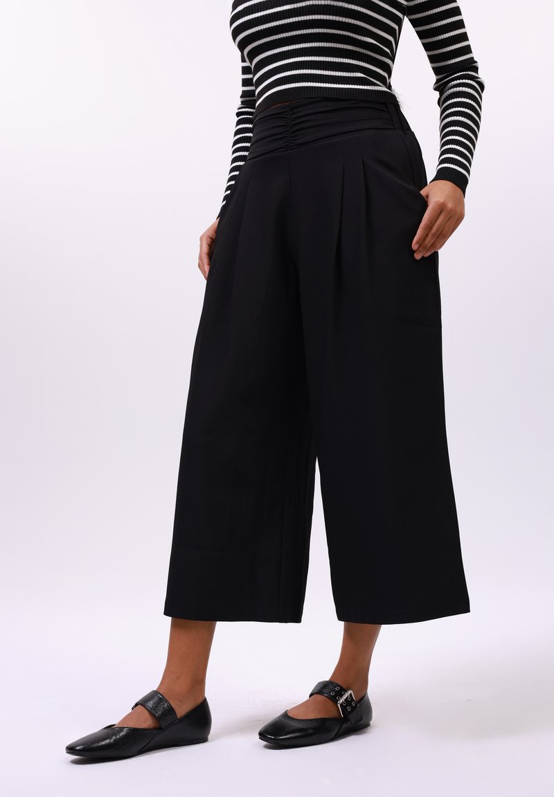 Pantalons larges noirs avec plis, mettant en avant une taille haute froncée. Le modèle porte des ballerines noires avec un détail de boucle. Haut rayé horizontal.